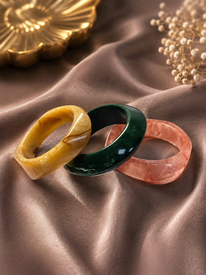 Three marble resin bangles in amber, green, and rose arranged overlapping on brown satin fabric