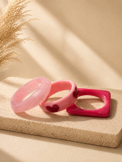 Three pink resin bangles in pale pink marbled, bubblegum pink with dark red hearts, and square hot pink, displayed on a stone surface