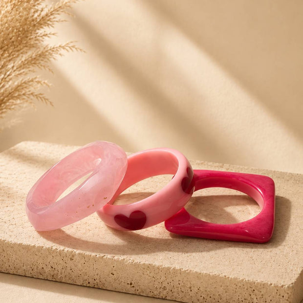 Three pink resin bangles in pale pink marbled, bubblegum pink with dark red hearts, and square hot pink, displayed on a stone surface