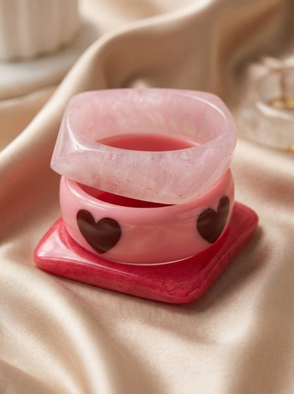 Close-up of three stacked pink resin bangles showing heart motifs and glossy finish, arranged on a hot pink square bangle base on satin fabric