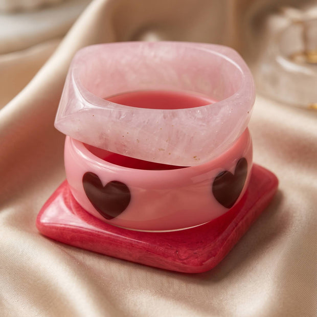 Close-up of three stacked pink resin bangles showing heart motifs and glossy finish, arranged on a hot pink square bangle base on satin fabric
