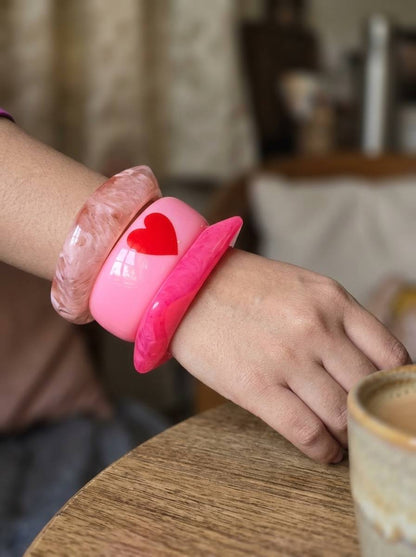 Pink resin bangles stacked on a woman's wrist resting on a wooden table beside a ceramic coffee cup