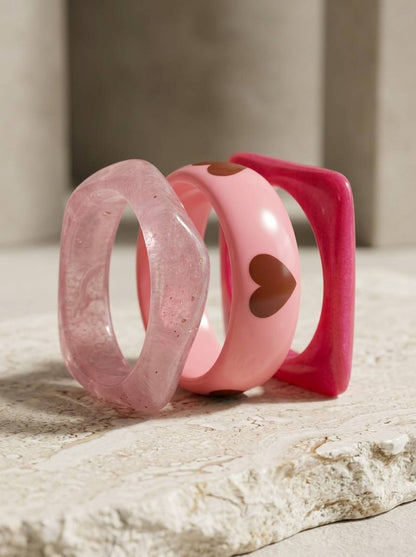 Three pink resin bangles standing upright on a stone surface showing translucent marbled, heart detail, and square silhouette styles