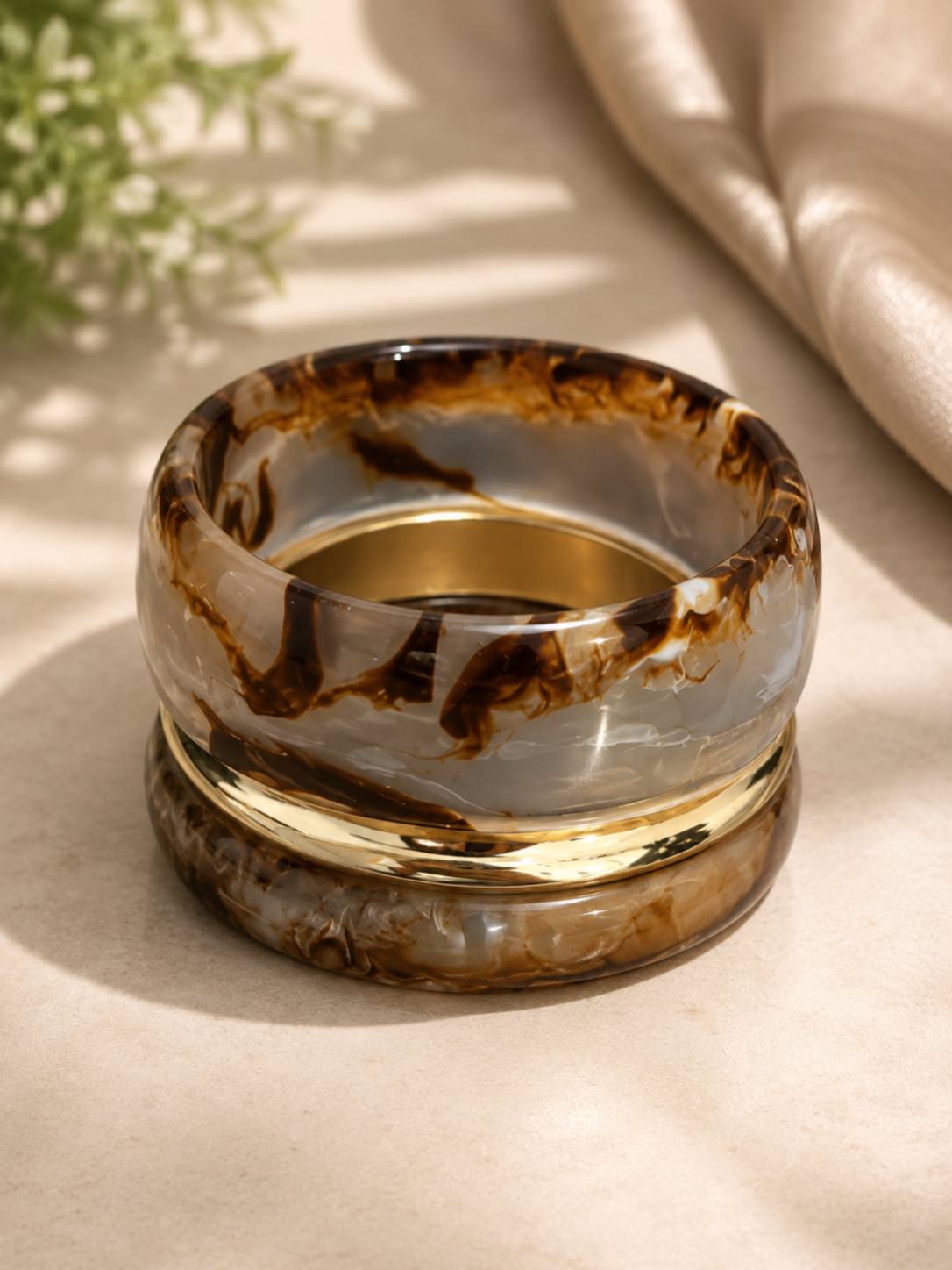 Three stacked bangles — two translucent brown marble resin and one gold-tone kada — resting on a beige surface