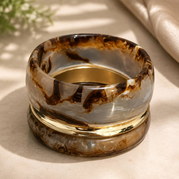 Three stacked bangles — two translucent brown marble resin and one gold-tone kada — resting on a beige surface