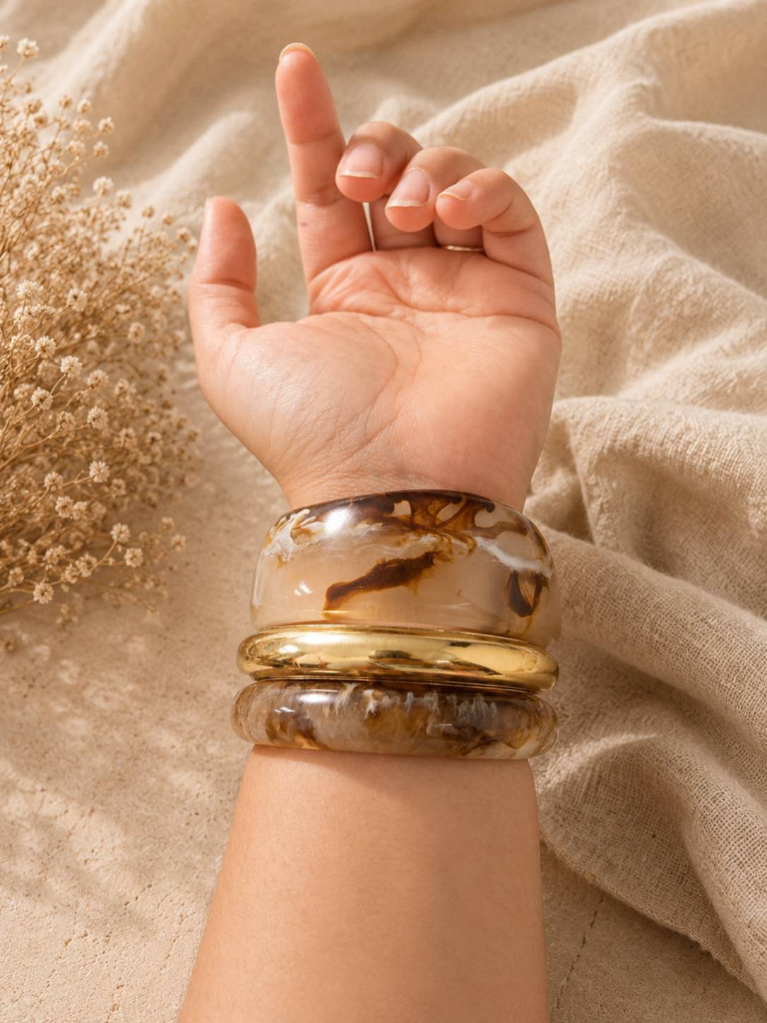 Resin marble bangles and gold-tone kada stacked on a woman's wrist raised against a beige linen backdrop