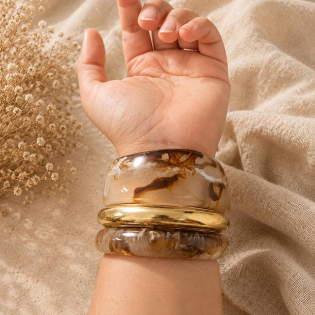 Resin marble bangles and gold-tone kada stacked on a woman's wrist raised against a beige linen backdrop