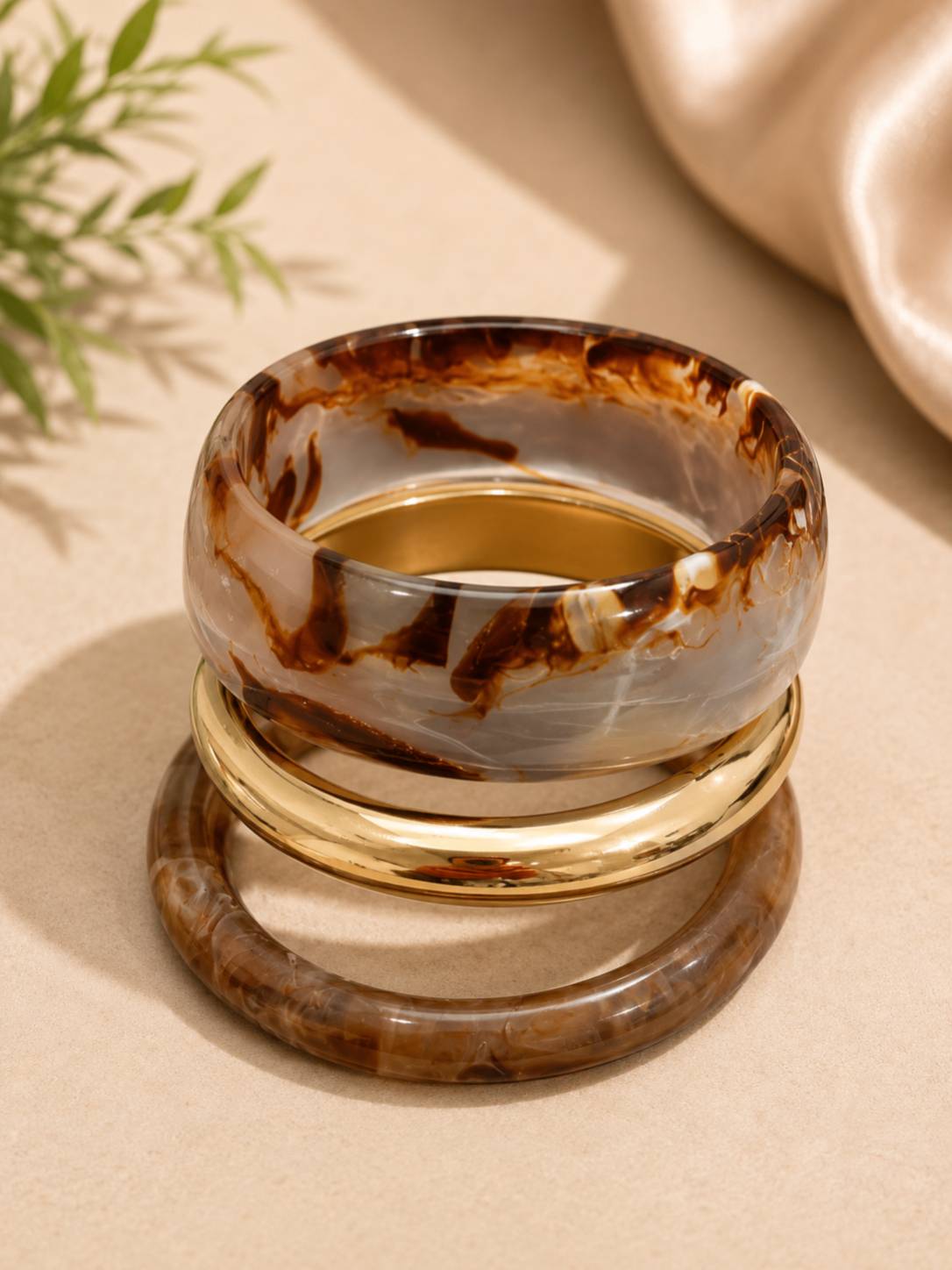 Side view of two marble resin bangles and a shiny gold-tone kada stacked together on a beige surface
