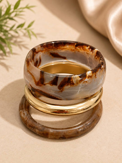Side view of two marble resin bangles and a shiny gold-tone kada stacked together on a beige surface