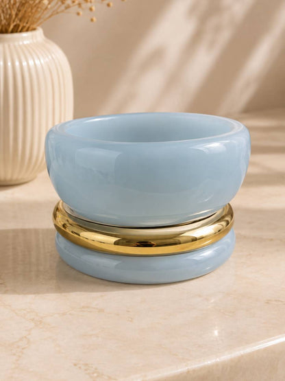 Three stacked bangles — two glossy light blue resin and one gold-tone metal — resting on a marble surface beside a white ribbed vase