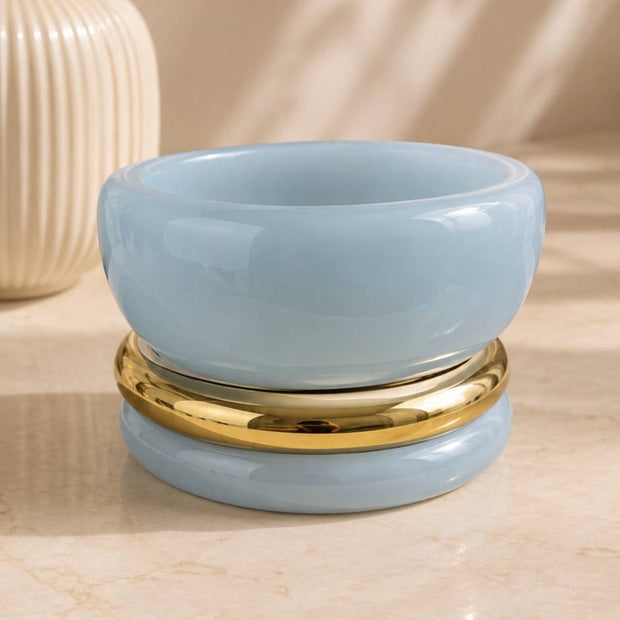 Three stacked bangles — two glossy light blue resin and one gold-tone metal — resting on a marble surface beside a white ribbed vase