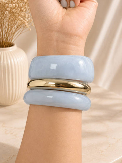 Three stacked bangles worn on a wrist — two chunky light blue resin bangles with a gold-tone metal bangle between them