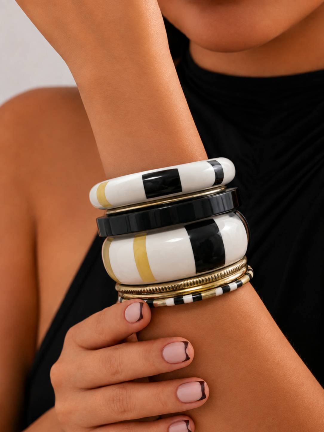 Woman wearing stacked black white and gold enamel bangles on her wrist against a black top with French-tip nails