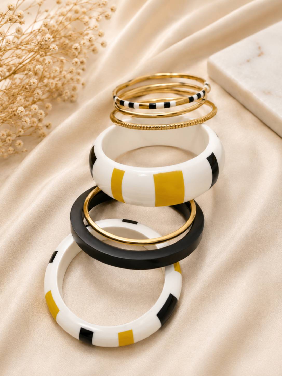 Stacked black white mustard yellow and gold-tone enamel bangles displayed on draped cream satin fabric