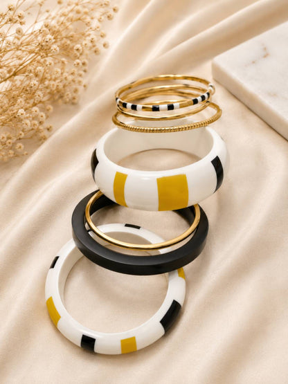 Stacked black white mustard yellow and gold-tone enamel bangles displayed on draped cream satin fabric