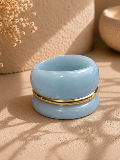 Three stacked blue marble resin bangles with gold-tone ring resting on sandy beige surface