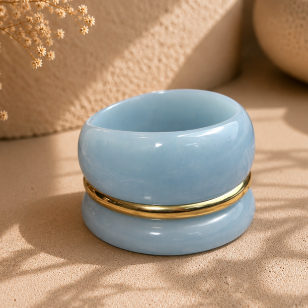 Three stacked blue marble resin bangles with gold-tone ring resting on sandy beige surface