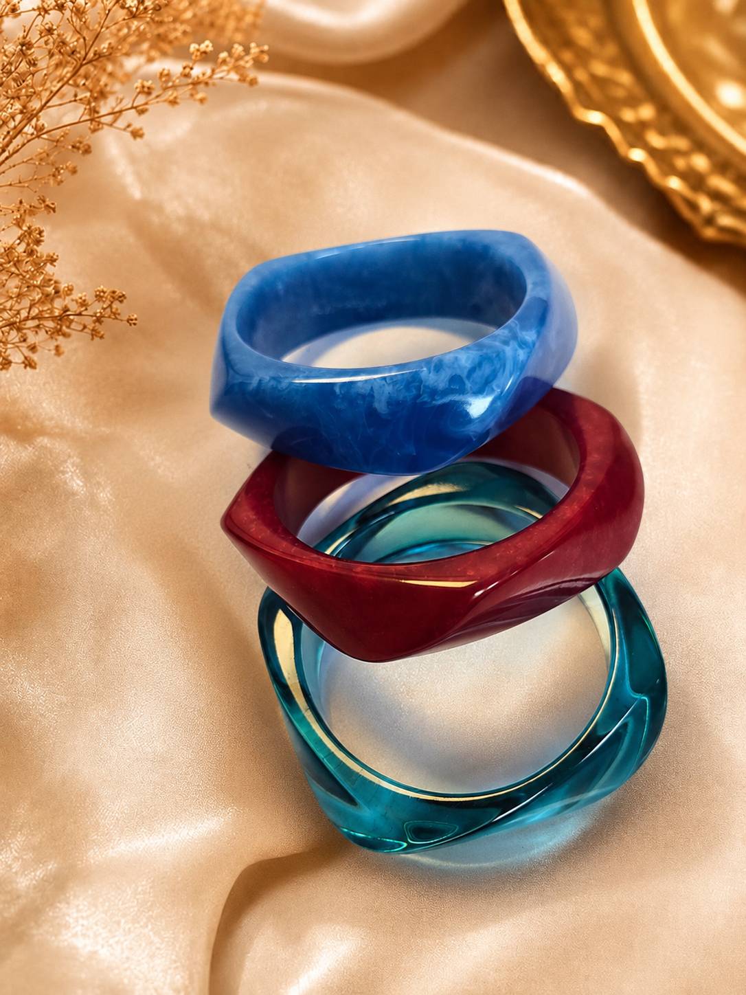 Three stacked resin marble bangles in blue, burgundy and teal on a satin cloth with dried flowers