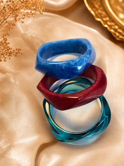 Three stacked resin marble bangles in blue, burgundy and teal on a satin cloth with dried flowers