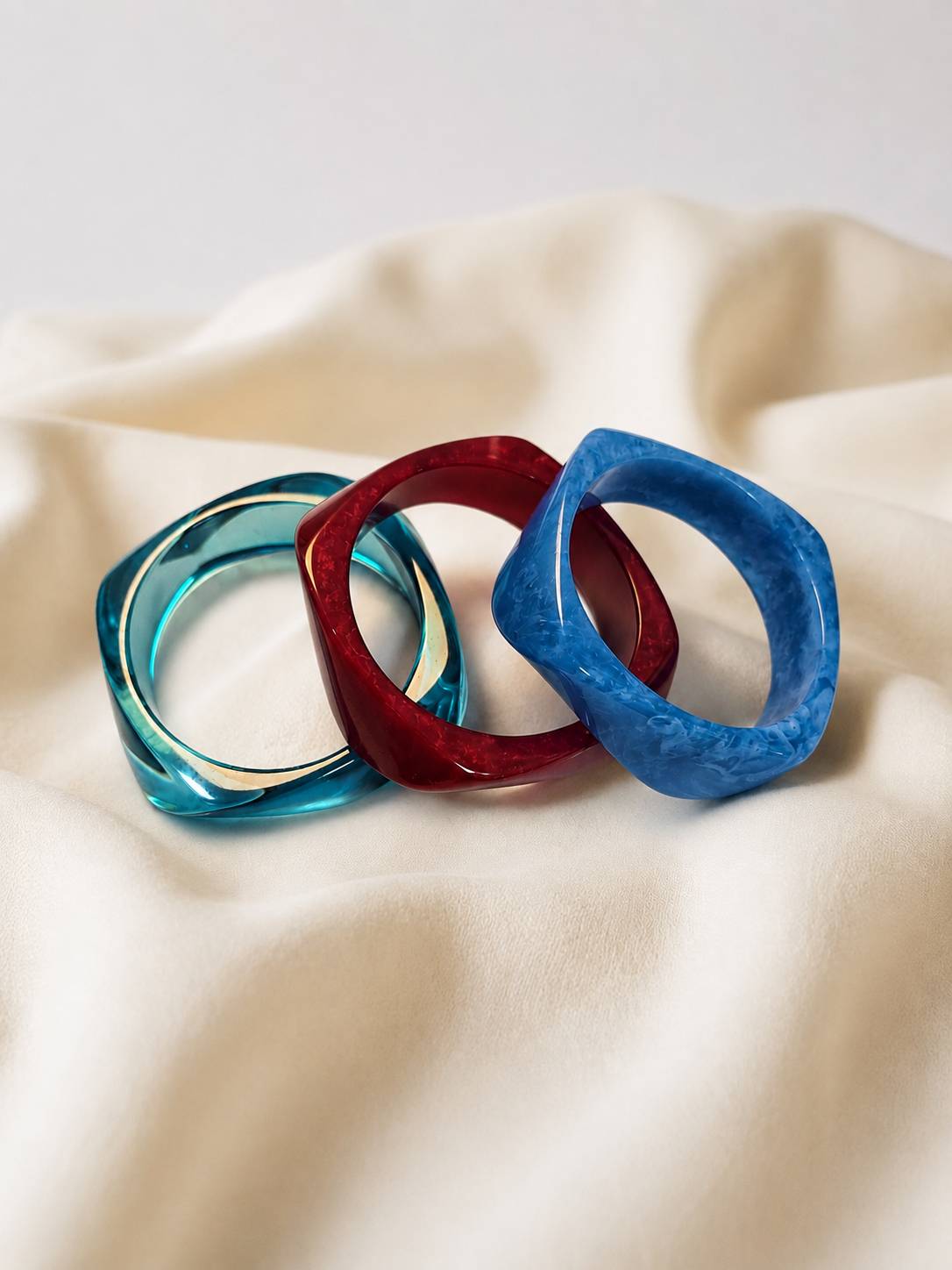 Cobalt blue, deep red and teal resin bangles arranged side by side on cream draped fabric