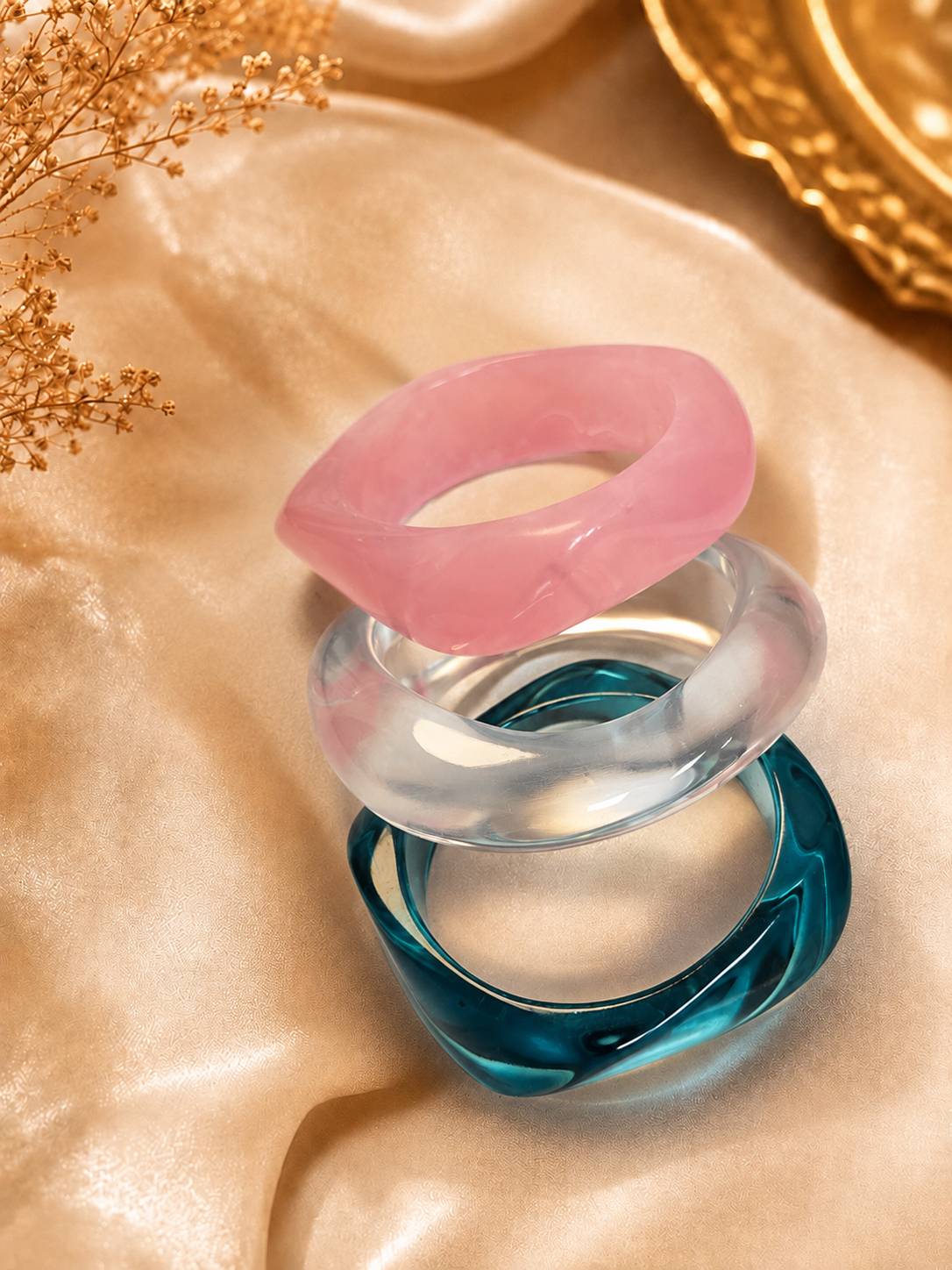 Three stacked resin bangles in pink, clear and teal on golden satin fabric with dried florals