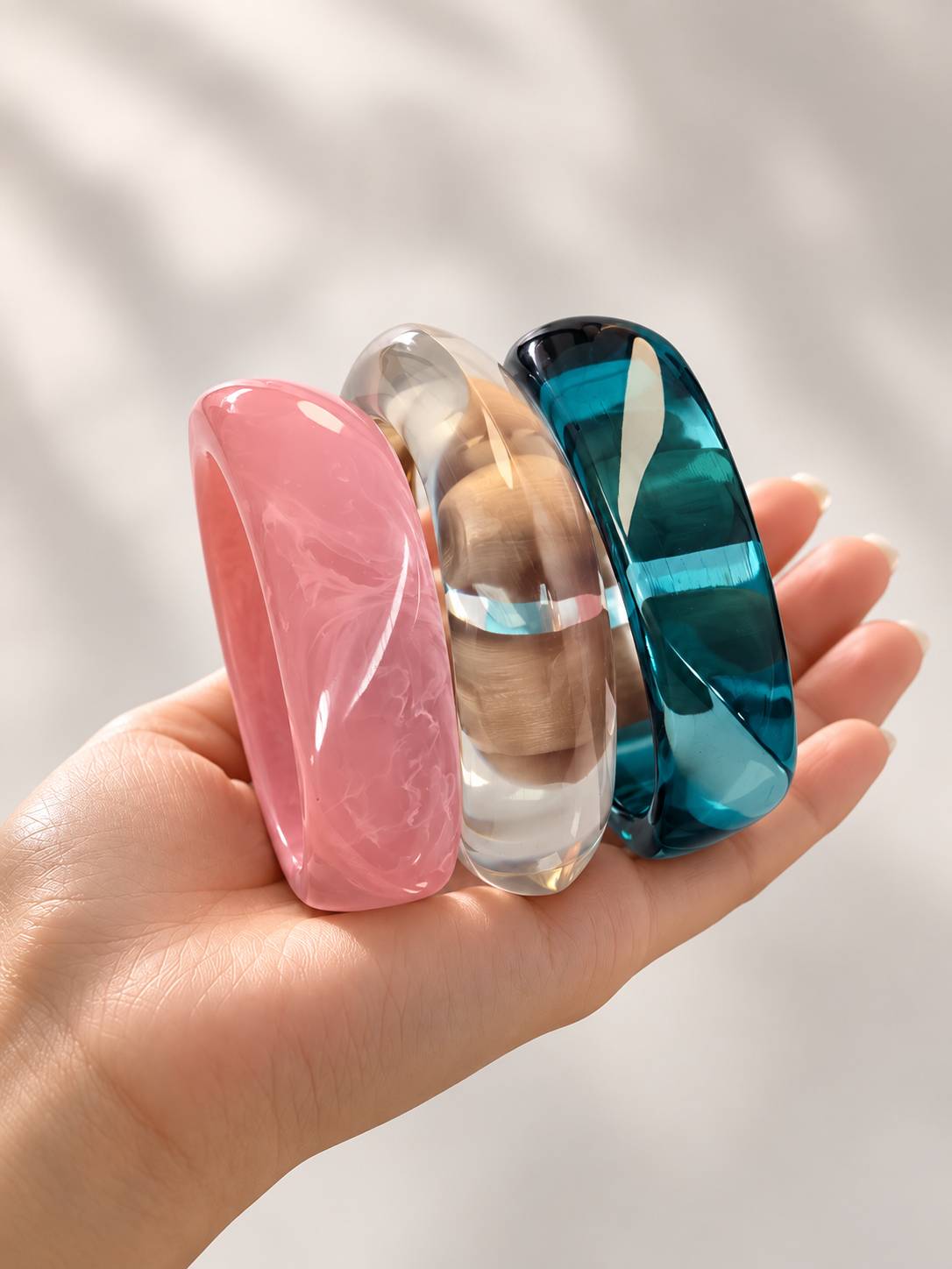 Hand holding three chunky resin bangles in pink, clear and teal upright against white background