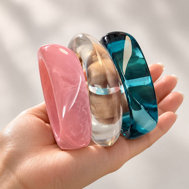 Hand holding three chunky resin bangles in pink, clear and teal upright against white background