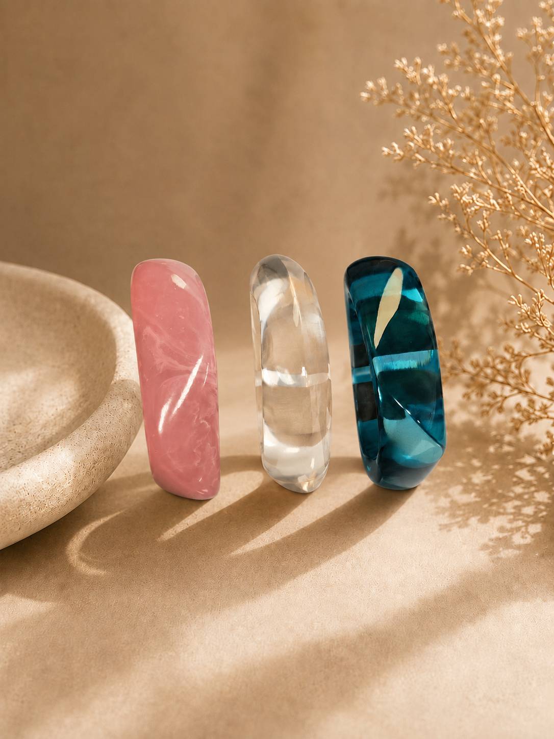 Pink, clear and teal resin bangles standing upright beside a stone bowl and dried flowers