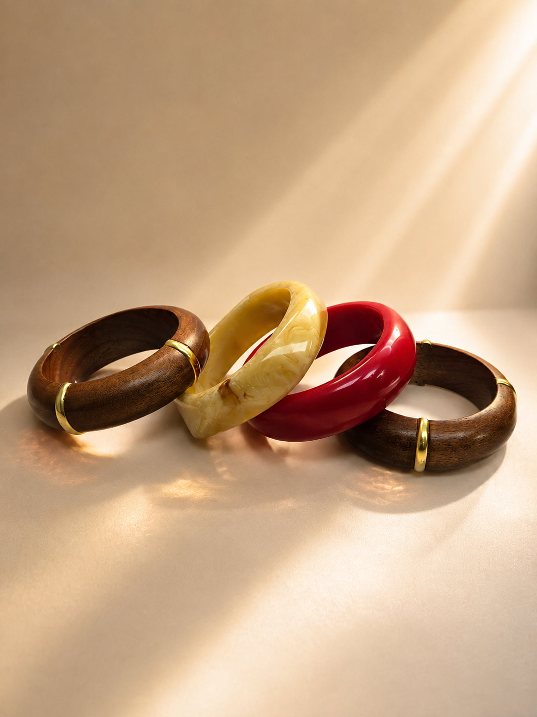 Four bangles laid flat in sunlight — two wood with gold accents, one cream marble resin, one red glossy resin