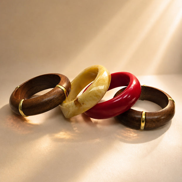 Four bangles laid flat in sunlight — two wood with gold accents, one cream marble resin, one red glossy resin