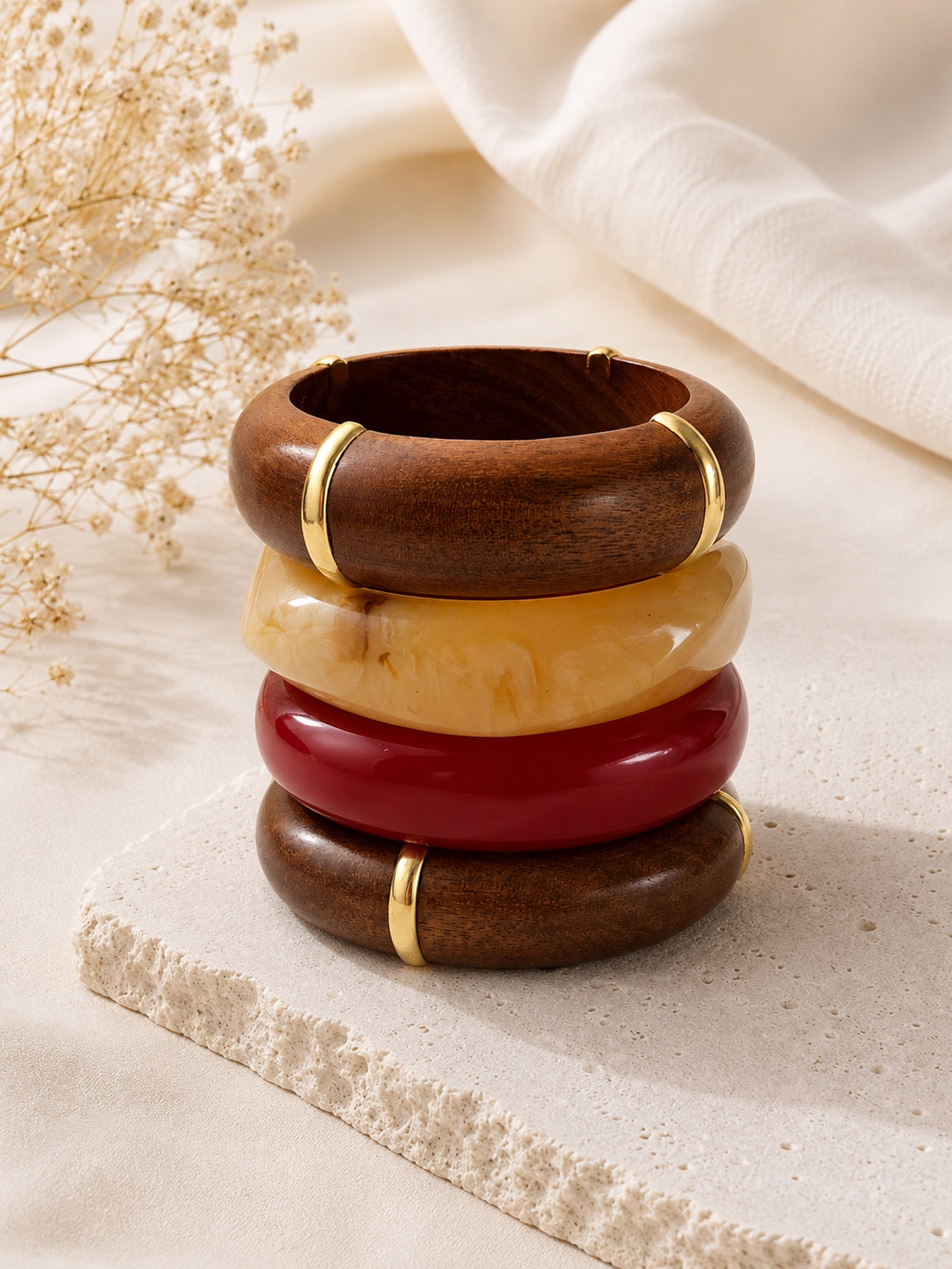 Four bangles stacked vertically on a stone surface beside dried flowers and white fabric