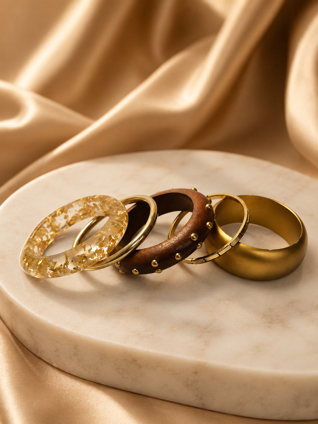 Four gold-tone bangles laid flat on a marble disc against draped gold satin — resin, metal, wood, and matte cuff