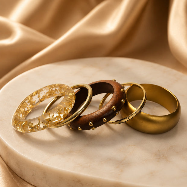 Four gold-tone bangles laid flat on a marble disc against draped gold satin — resin, metal, wood, and matte cuff