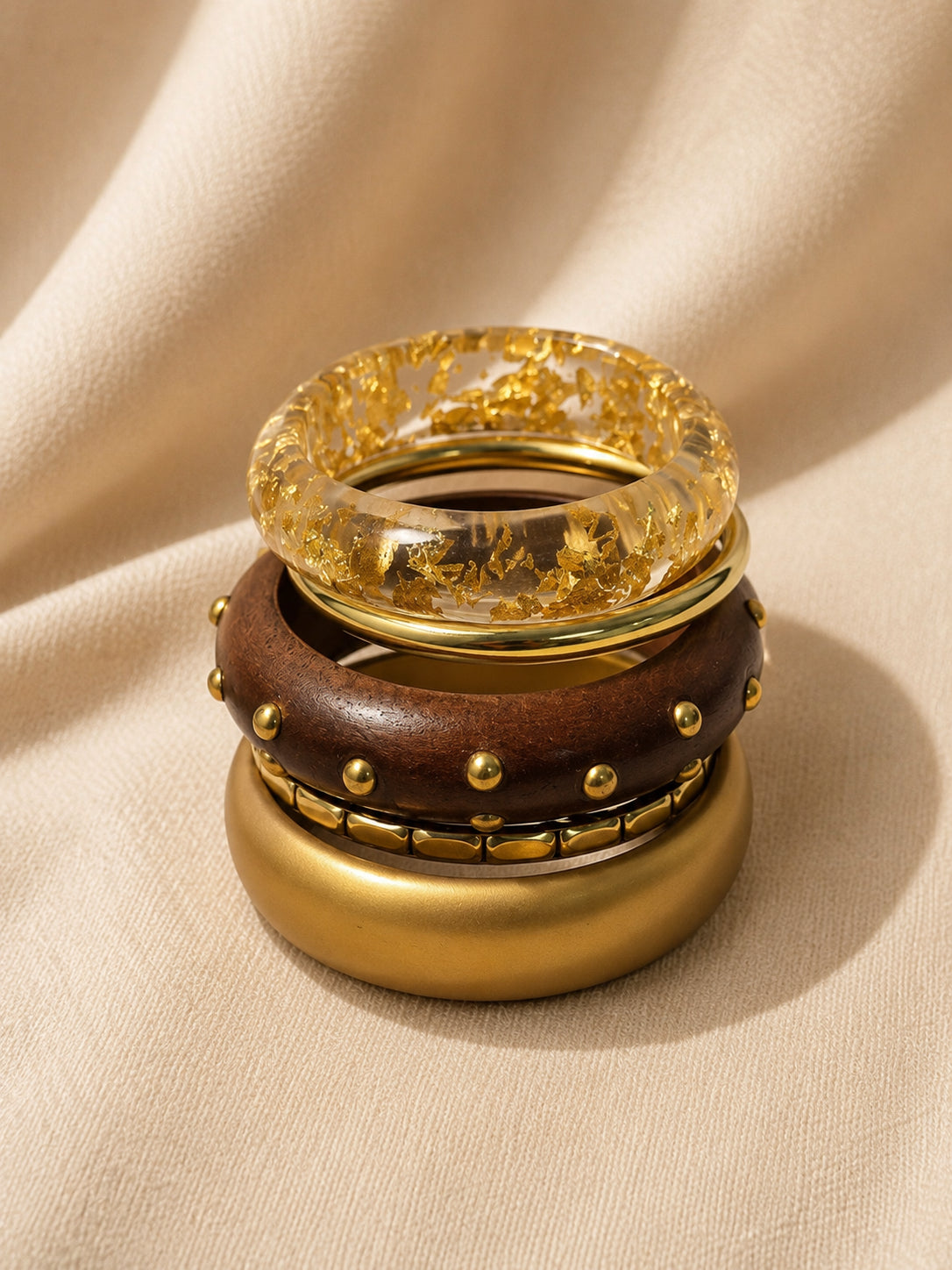 Four stacked bangles piled vertically showing resin with gold foil on top, studded wood bangle in the middle, and matte gold cuff at the base