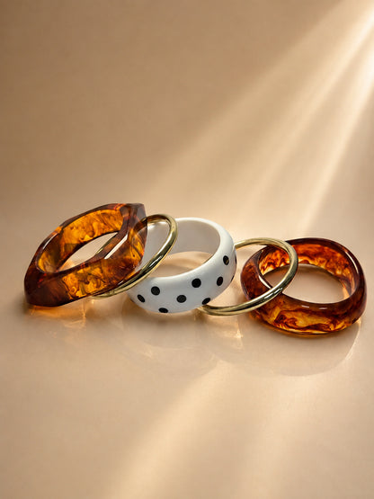 Five bangles laid flat showing two tortoiseshell resin cuffs, one angular resin bangle, two gold-tone rings, and a white polka-dot bangle on a beige surface