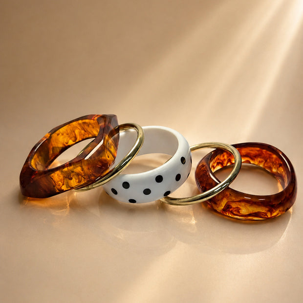 Five bangles laid flat showing two tortoiseshell resin cuffs, one angular resin bangle, two gold-tone rings, and a white polka-dot bangle on a beige surface