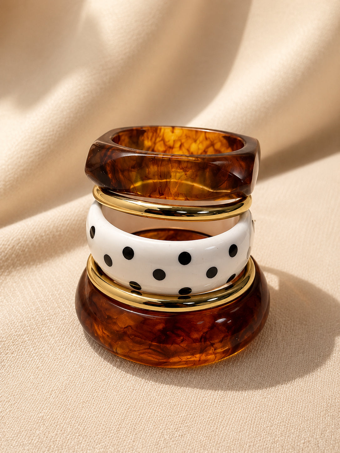 Tortoiseshell resin bangles stacked vertically with white polka-dot bangle and gold-tone rings on cream fabric background