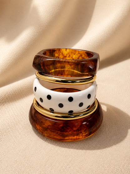 Tortoiseshell resin bangles stacked vertically with white polka-dot bangle and gold-tone rings on cream fabric background