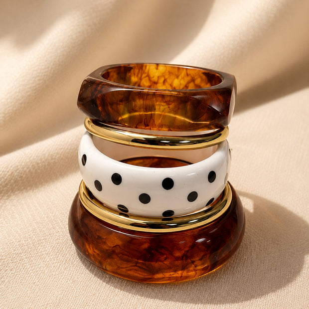 Tortoiseshell resin bangles stacked vertically with white polka-dot bangle and gold-tone rings on cream fabric background