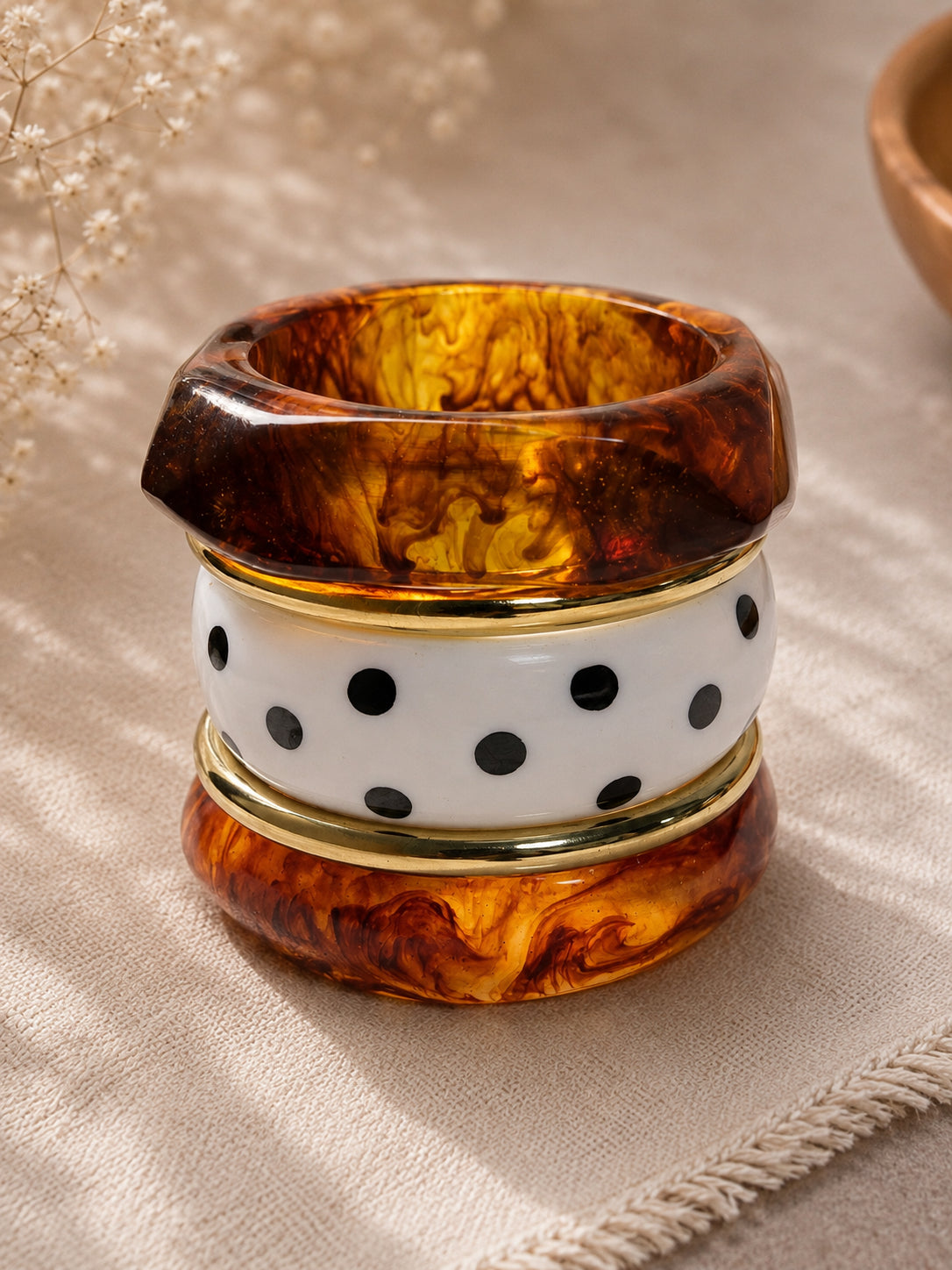 Close-up stack of angular tortoiseshell resin bangle, white polka-dot enamel bangle, gold-tone rings, and round resin cuff on linen fabric