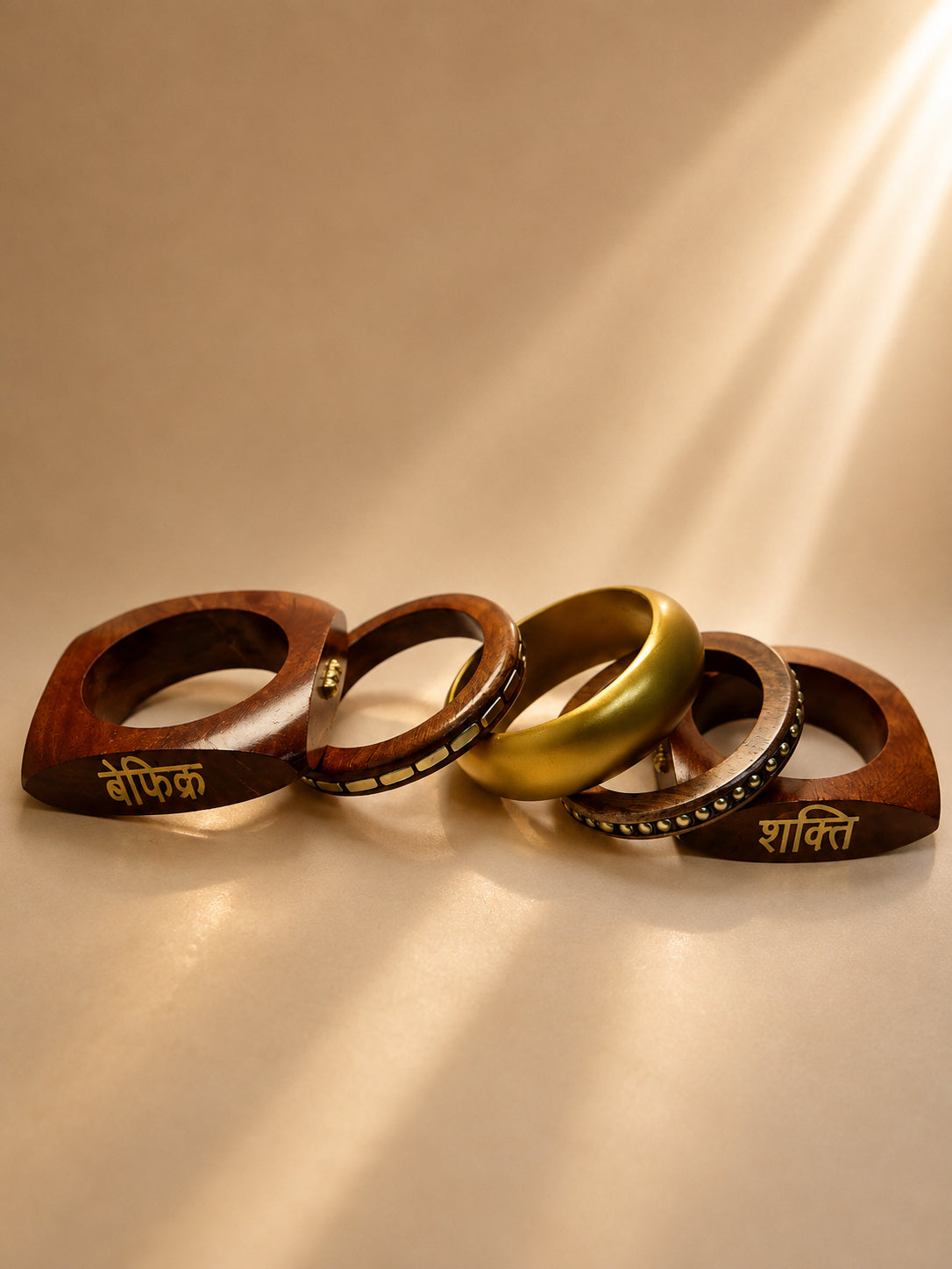 Five stacked bangles laid flat showing two geometric wood bangles engraved in Hindi and three gold-toned metal bangles with sunlight streaming across them