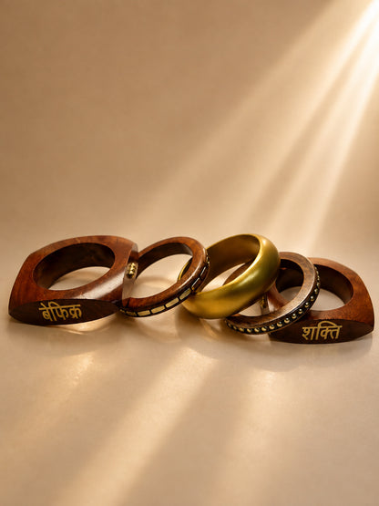 Five stacked bangles laid flat showing two geometric wood bangles engraved in Hindi and three gold-toned metal bangles with sunlight streaming across them