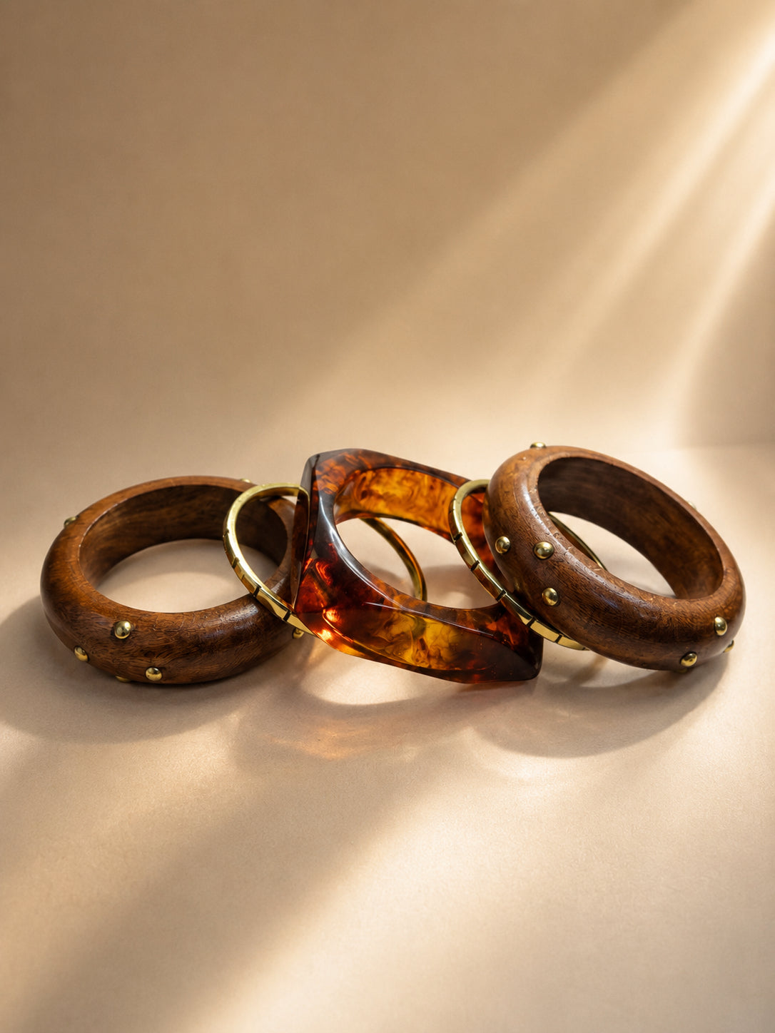 Three stacked bangles laid flat — two wood bangles with brass dome studs and one square tortoise resin cuff with gold inlay