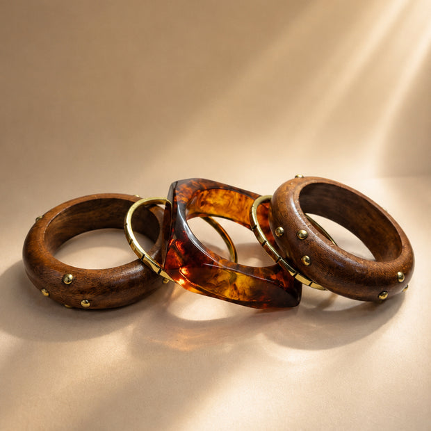 Three stacked bangles laid flat — two wood bangles with brass dome studs and one square tortoise resin cuff with gold inlay