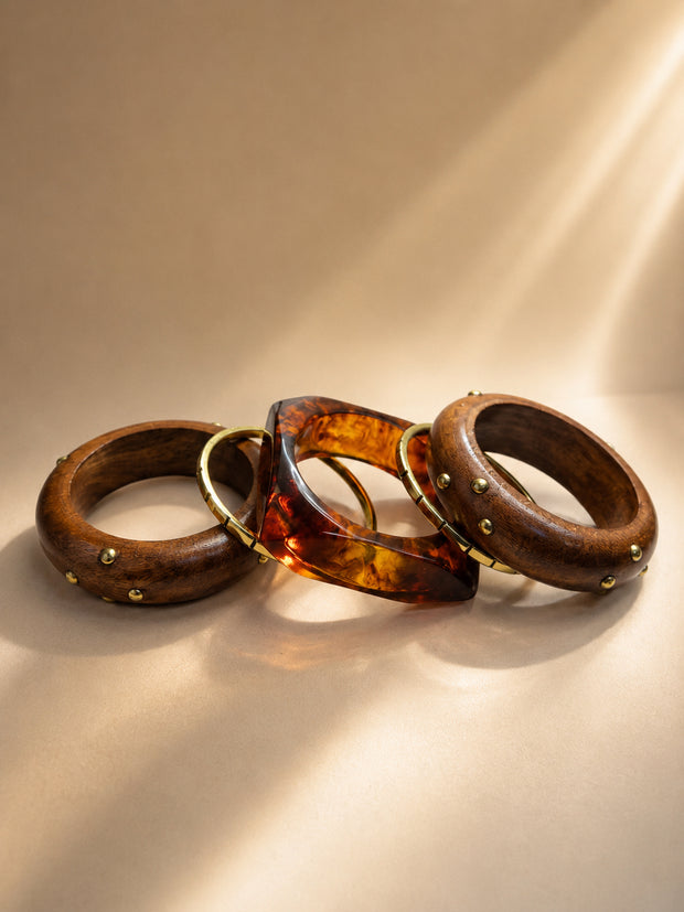 Three stacked bangles laid flat — two wood bangles with brass dome studs and one square tortoise resin cuff with gold inlay