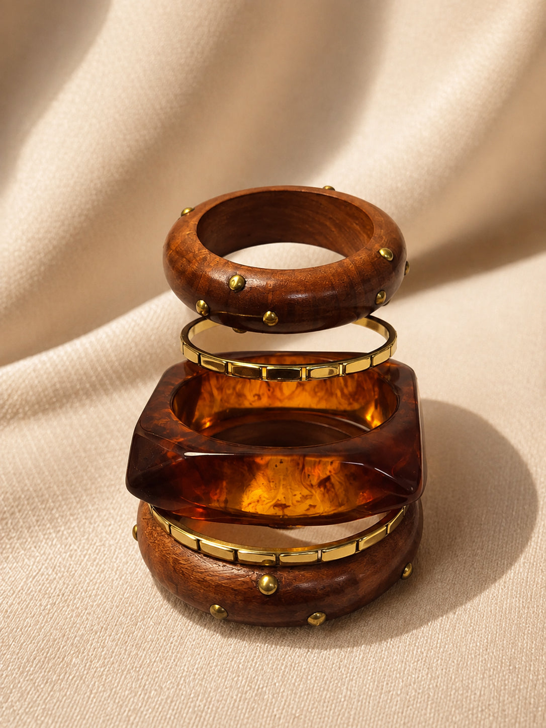 Four bangles stacked vertically against draped cream fabric, showing the square resin cuff sandwiched between two round wood bangles and a slim brass bangle
