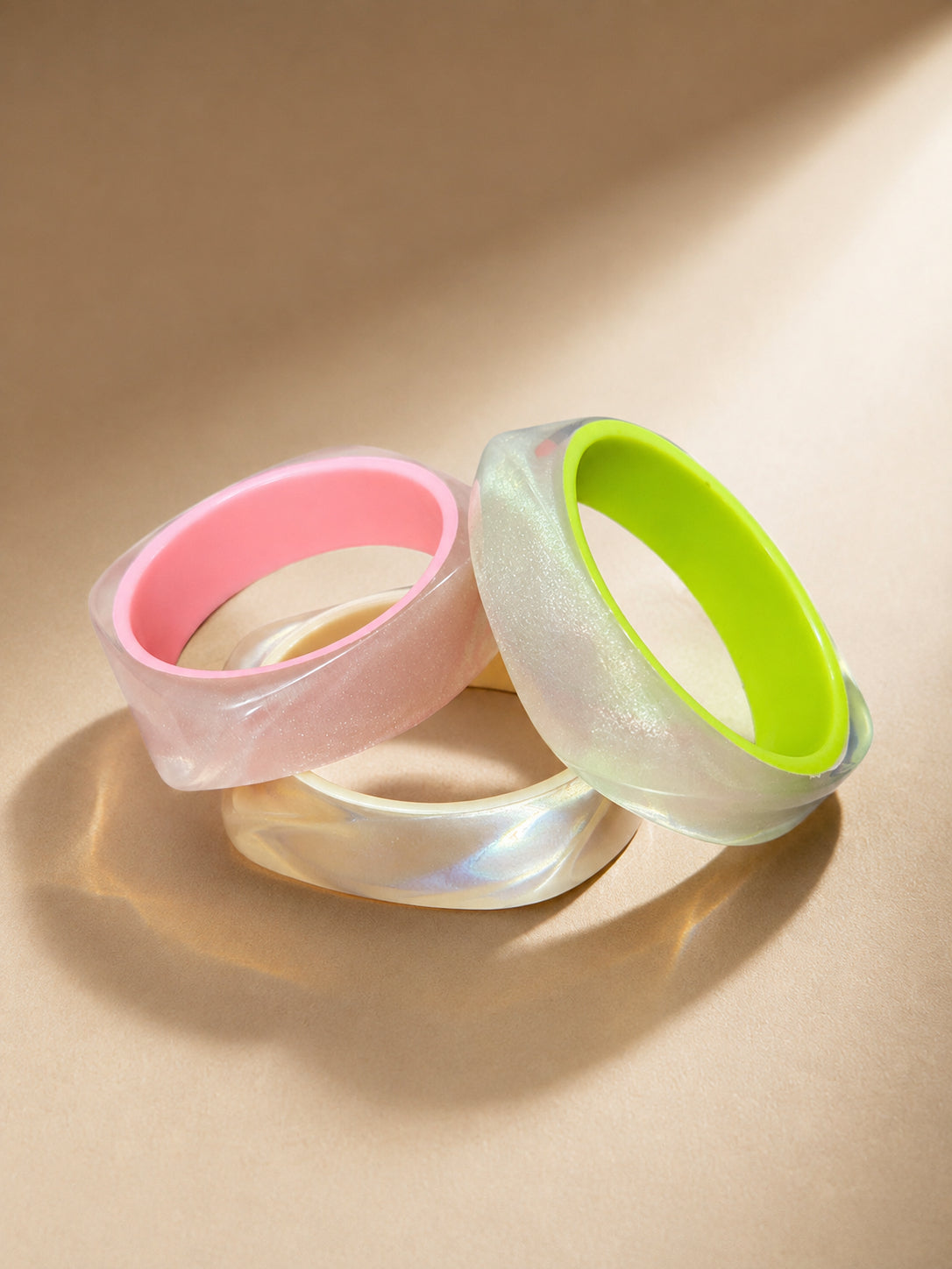 Three pearlescent resin bangles with pink and neon green inner rims arranged on a beige surface