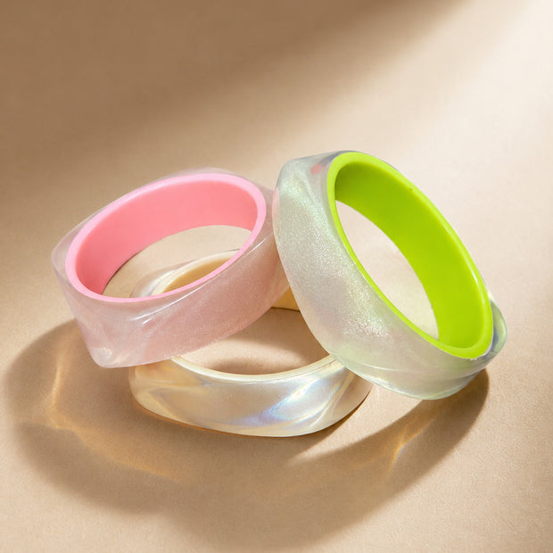 Three pearlescent resin bangles with pink and neon green inner rims arranged on a beige surface
