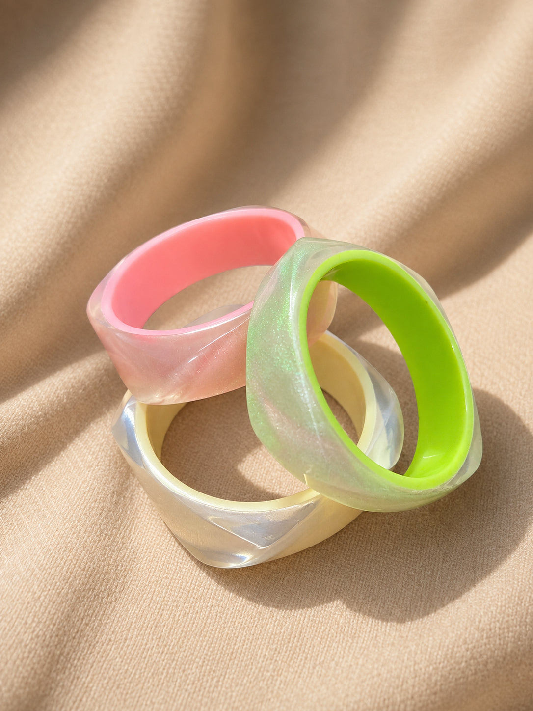 Three faceted pearlescent resin bangles in ivory, pink, and neon green laid on draped beige fabric
