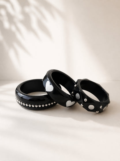 Three black resin bangles with silver studs arranged in a fan on a white surface with leaf shadow